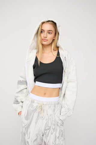 Woman wearing a black sports bra and gray sweatpants with a white jacket on a light gray background