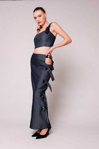 A model poses in a dark navy blue two-piece outfit consisting of a halter crop top with side bows and a matching high-waisted maxi skirt with side cutouts and hanging bows, set against a plain light gray studio backdrop.