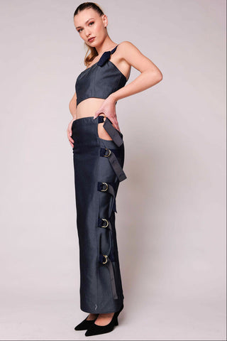 A model stands against a light gray studio backdrop wearing a dark navy two-piece outfit consisting of a halter crop top and matching wide-leg pants with side cutouts and buckle details, paired with black pointed shoes.