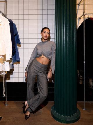 Woman in a gray outfit standing in a clothing store with a green column and rack of clothes in the background.
