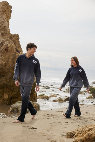 Models walking along beach wearing unisex hoodie offering a cozy fit, featuring an Aries zodiac symbol on the chest, "Aries" on the right arm, Aries constellation on the back, and the words "We collide, shattering my mind," made with sustainable, luxurious materials